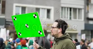 年轻的独立男子拿着绿色<strong>屏</strong>幕的<strong>大海报</strong>，在城市集会上的人群中行走在街道上，侧视。